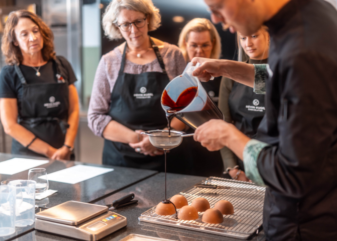 Der Patissier-Chef zeigt den Teilnehmern wie die Glasur über Törtchen gegossen wird. In der Hand hält er die Glasur und ein Sieb.